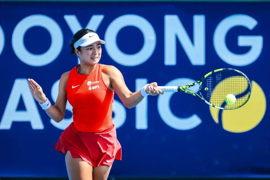 A tennis player in a red sleeveless top and red skirt with a white visor, swinging a forehand at a ball during a match at the Dubai Duty Free... no, the sign…