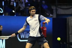 Tennis player in white and navy EA7 shirt and shorts hitting a forehand near a glowing ball at an indoor match; Alexander Bublik