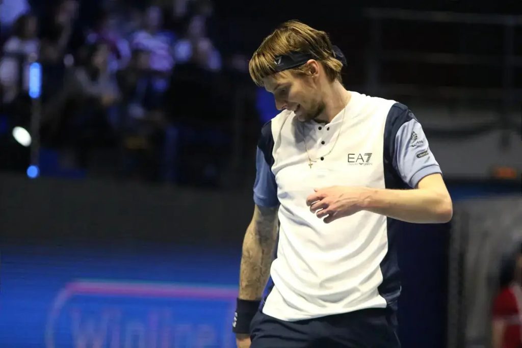 Tennis player in white EA7 shirt and black headband smiling while walking on court during a match, with blurred crowd behind.