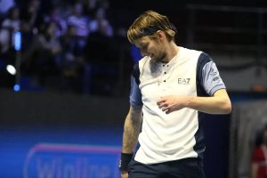 Tennis player in white EA7 shirt and black headband smiling while walking on court during a match, with blurred crowd behind.