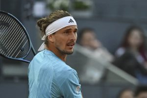 Tennis player in a light blue shirt and white Adidas headband, holding a racquet on a clay court, looking to the side during a match