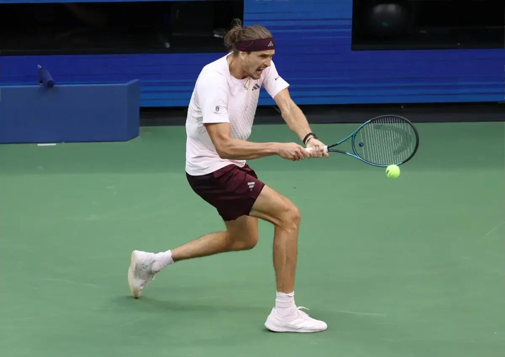 Tennis player in a pink shirt and maroon shorts hits a forehand on a green court, with a blue racket and yellow ball in play.