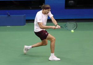 Tennis player in a pink shirt and maroon shorts hits a forehand on a green court, with a blue racket and yellow ball in play.