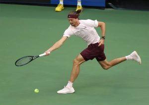 Tennis player in maroon shorts and white shirt lunging to return a forehand on a green court, yellow ball near his feet. Story about Zverev and the Laver Cup.