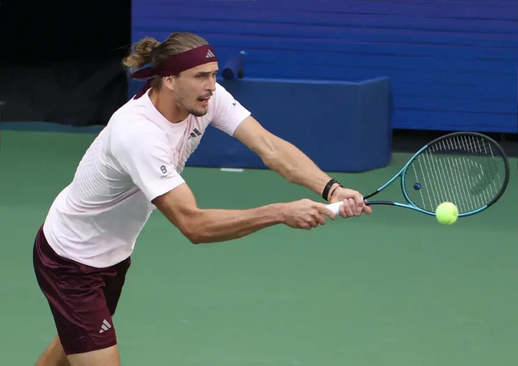 A tennis player in a white shirt and maroon shorts returns a forehand with a blue racket on a green court.