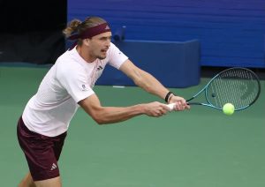 A tennis player in a white shirt and maroon shorts returns a forehand with a blue racket on a green court.