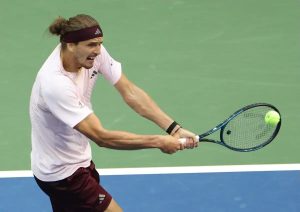 Tennis player in a maroon headband and white Adidas shirt hitting a backhand on a green court, ball visible near racket