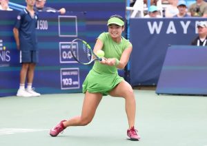 Tennis player in a green dress and visor hitting a forehand on a hard court, with "Tennis" and "Wayne" signage behind her. The article is about Alexandra Eal…