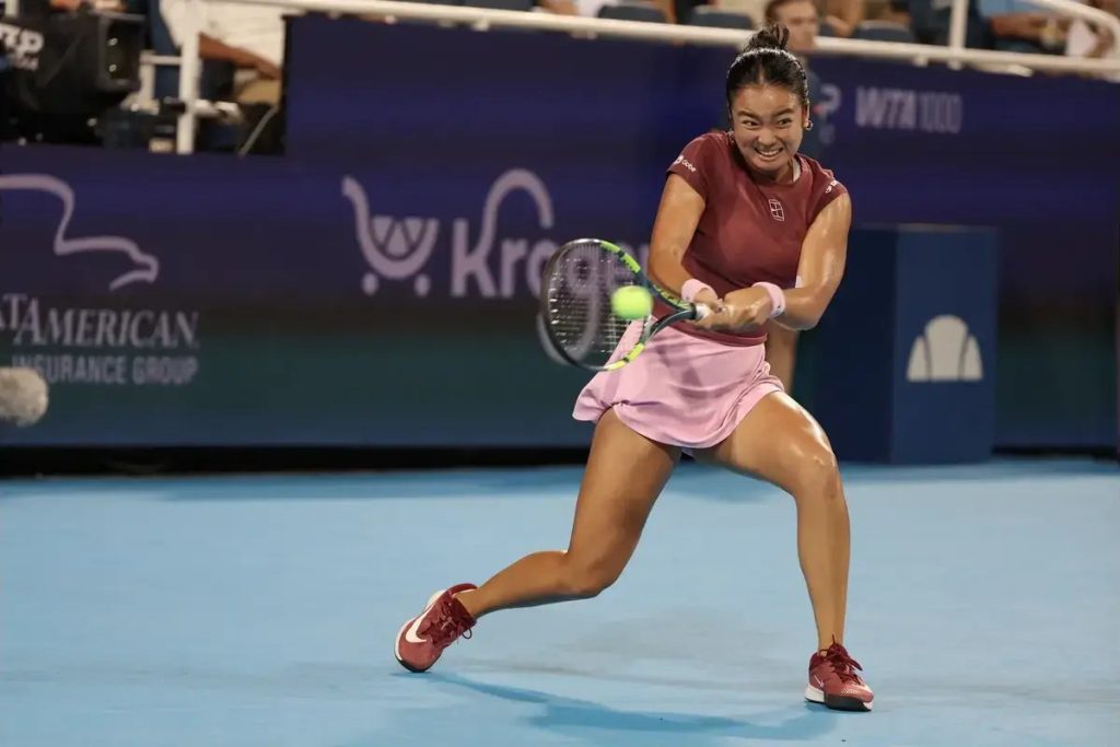 A tennis player in a maroon top and pink skirt swings a forehand on a blue court during a night match, with "WTA 1000" signage behind her.