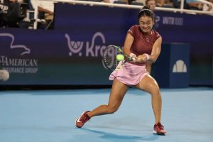 A tennis player in a maroon top and pink skirt swings a forehand on a blue court during a night match, with "WTA 1000" signage behind her.