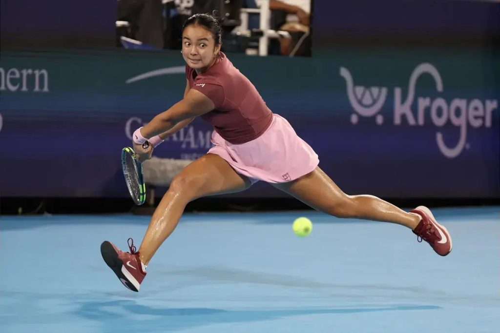 Tennis player in a maroon top and pink skirt lunging to hit a low forehand on a blue court, with a Kroger sign behind her. Article subject: Alexandra Eala.