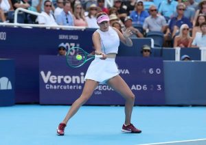 Tennis player in white Nike outfit and pink visor hitting a forehand on a blue court at a tournament; scoreboard shows "Veronique" sponsor and "USA" player n…