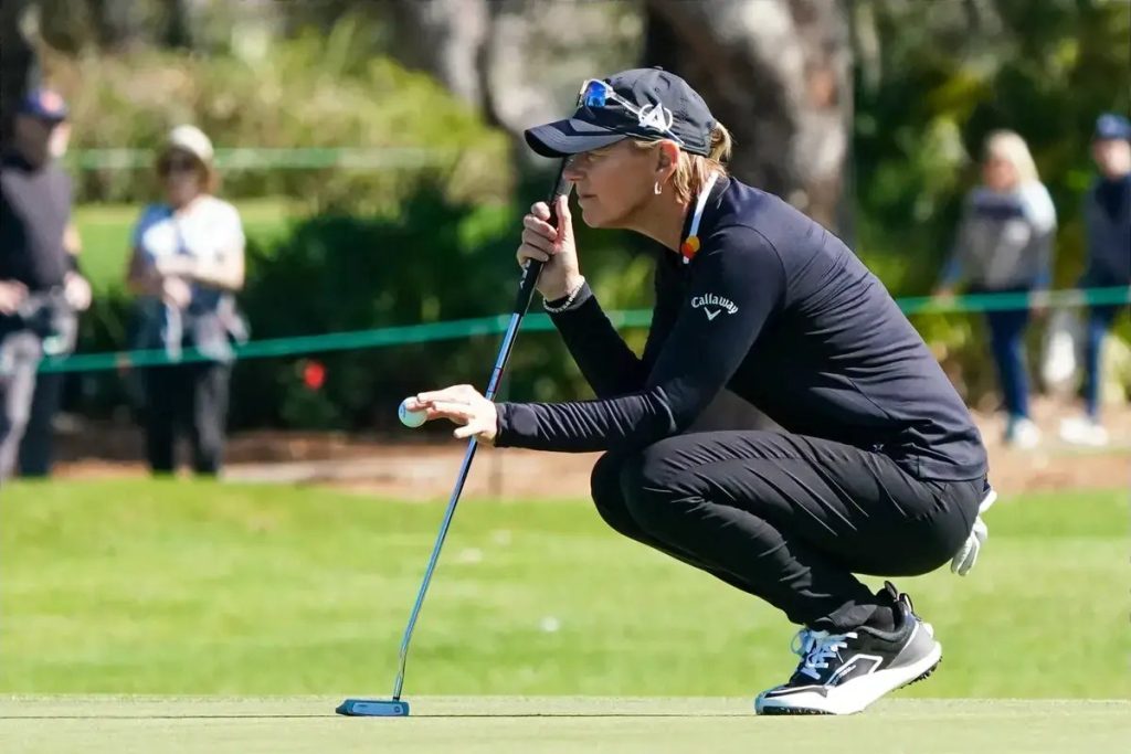 Golfer in black Callaway cap and long-sleeve top crouches to read a putt on the green, putter in hand, spectators behind a rope