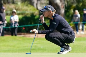 Golfer in black Callaway cap and long-sleeve top crouches to read a putt on the green, putter in hand, spectators behind a rope