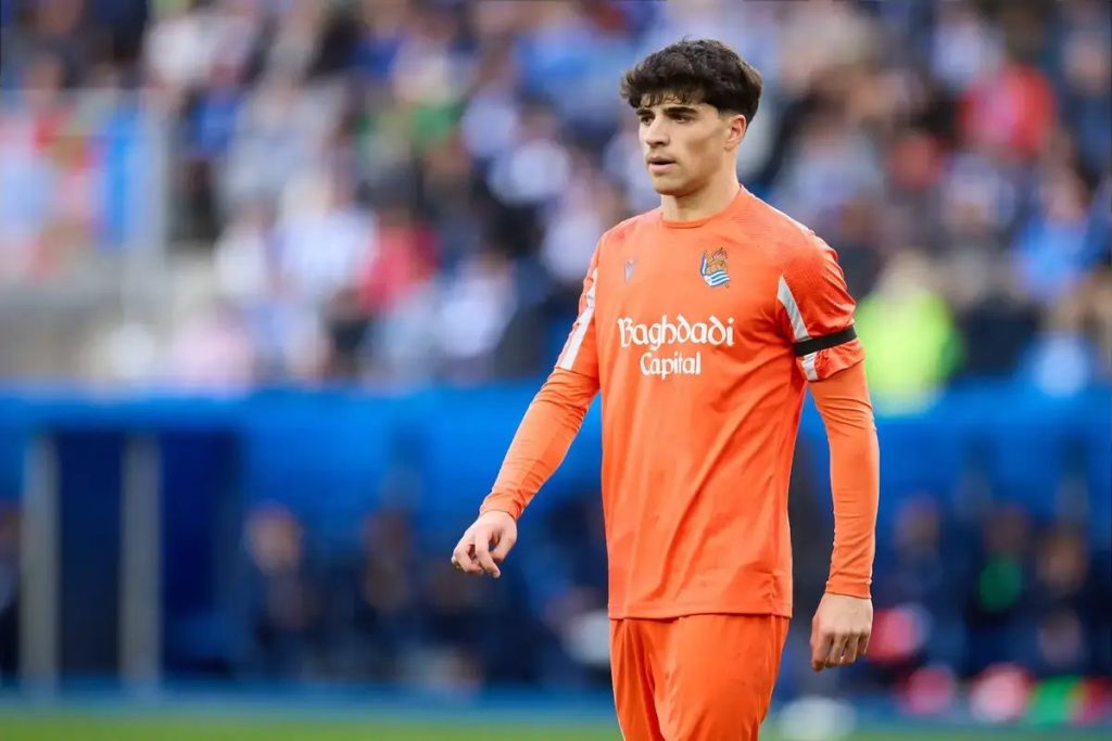Football player in orange Real Sociedad goalkeeper kit with Baghdadi Capital sponsor, standing on a pitch before a crowd