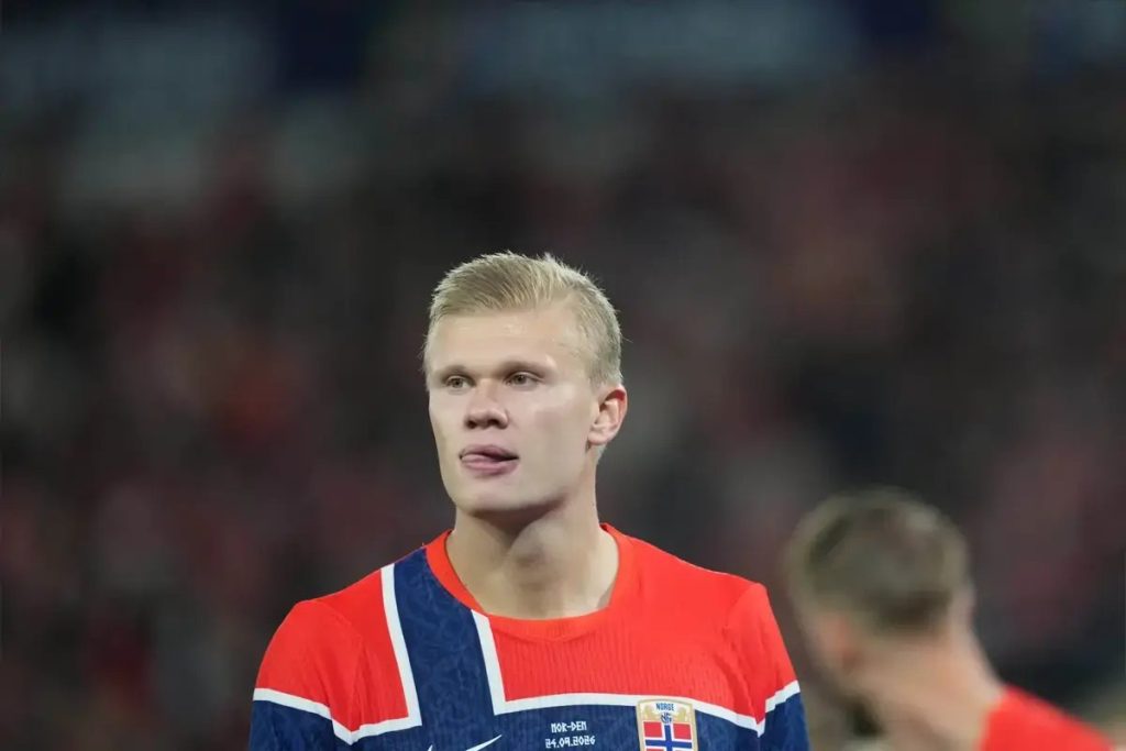 Footballer with blond hair in a red and navy Norway national team shirt looking ahead during a match, with a blurred crowd behind him.