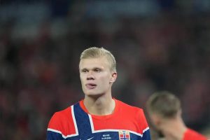 Footballer with blond hair in a red and navy Norway national team shirt looking ahead during a match, with a blurred crowd behind him.