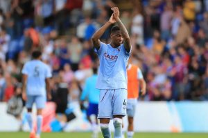 Player in light blue Aston Villa-style kit, number 4, applauding fans at a stadium, with a teammate wearing number 5 behind him