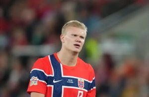 Blond footballer in red and blue Norway kit with national crest and Nations League badge, looking up during a match