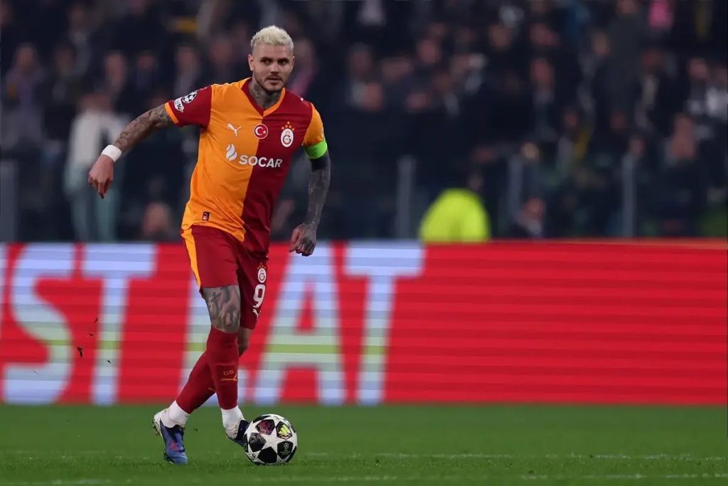 Footballer in orange and red Galatasaray kit, number 9, dribbling the ball on a pitch in a stadium at night.