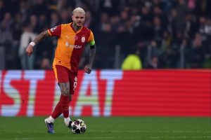 Footballer in orange and red Galatasaray kit, number 9, dribbling the ball on a pitch in a stadium at night.
