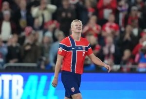 Football player in red and navy Norway kit, number 9, smiling and looking up at a packed stadium crowd during an international match.