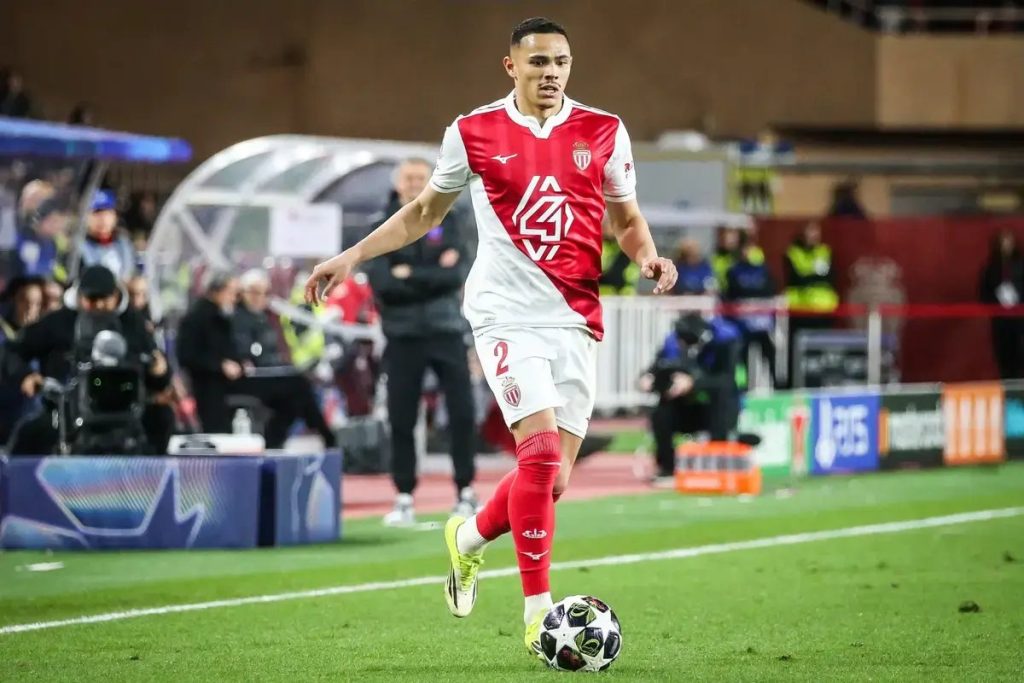 Monaco player wearing number 2 red and white kit dribbles the ball on the pitch during a match, with spectators and staff behind.