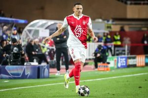 Monaco player wearing number 2 red and white kit dribbles the ball on the pitch during a match, with spectators and staff behind.