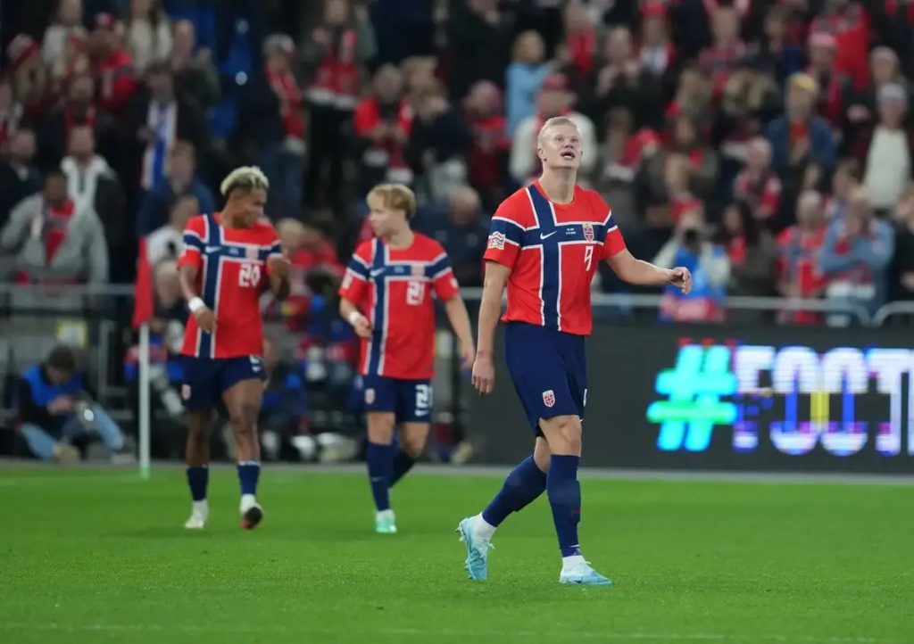 Football player in red and navy Norway kit with number 9 walking on pitch, looking up, teammates behind him in a stadium