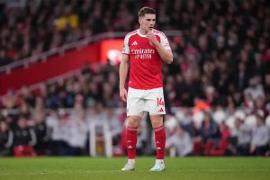 A footballer in red Arsenal home kit with number 14 and Emirates sponsor, touching his chin on the pitch during a match at a packed stadium