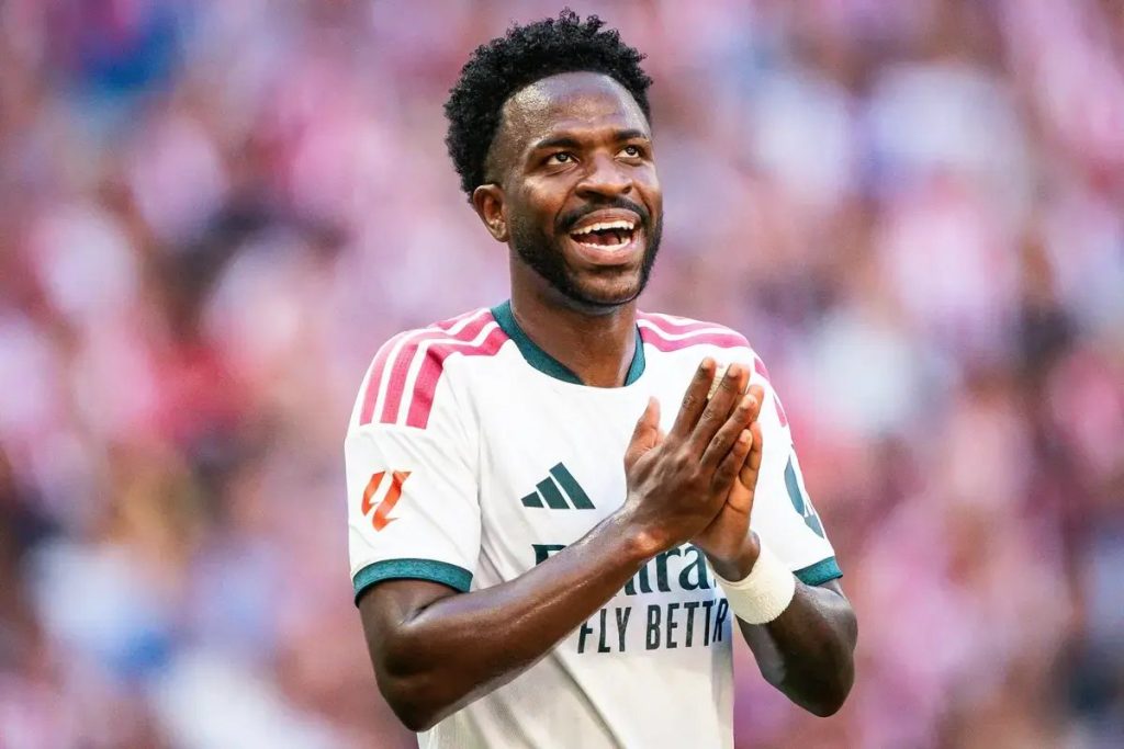 A smiling player in a white and teal Arsenal-style kit with "FLY BETTER" sponsor text, clapping hands together on a pitch.
