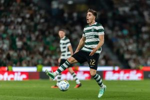 Sporting CP player number 22 in green-and-white stripes shouting and kicking the ball during a match at a packed stadium