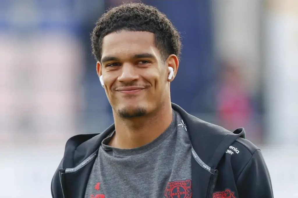 Young man with curly hair and white earbuds smiling, wearing a grey Bayer 04 Leverkusen shirt under a black zip jacket at a stadium.