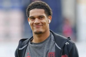 Young man with curly hair and white earbuds smiling, wearing a grey Bayer 04 Leverkusen shirt under a black zip jacket at a stadium.