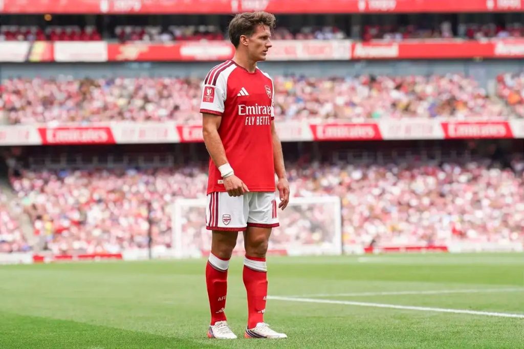Footballer in red and white Arsenal kit with Emirates sponsor, standing on the pitch at Emirates Stadium before a packed crowd