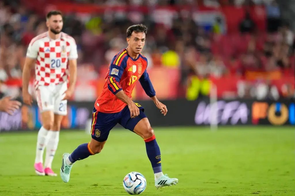 Spain midfielder in red and navy kit dribbling the ball past a Croatia player in a white checkerboard shirt during a match