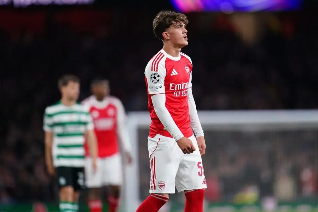 Young footballer in red and white Arsenal kit with Emirates sponsor and Champions League badge, looking up during a match, with Sporting player in green-and-…
