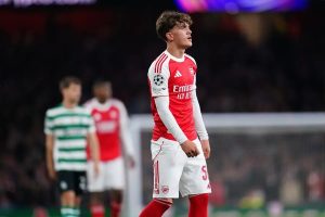 Young footballer in red and white Arsenal kit with Emirates sponsor and Champions League badge, looking up during a match, with Sporting player in green-and-…