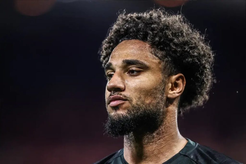 A footballer with curly hair and a beard, wearing a dark kit, looks off to the side in a dimly lit stadium setting