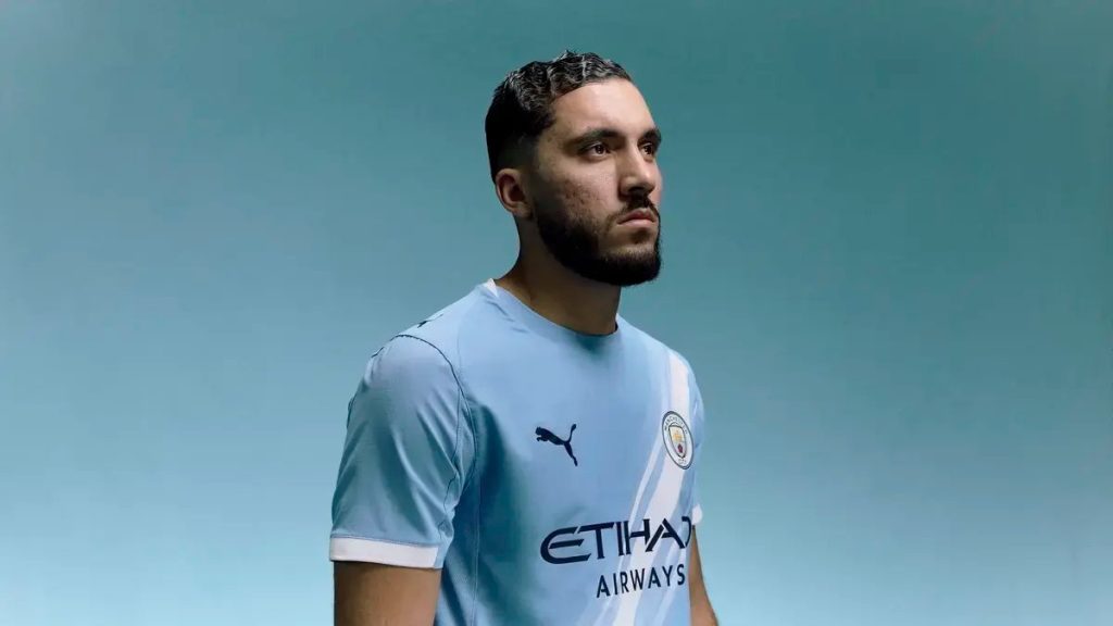 A bearded man with slicked-back dark hair in a light sky-blue Manchester City home shirt with Etihad Airways sponsor, posing against a blue backdrop.
