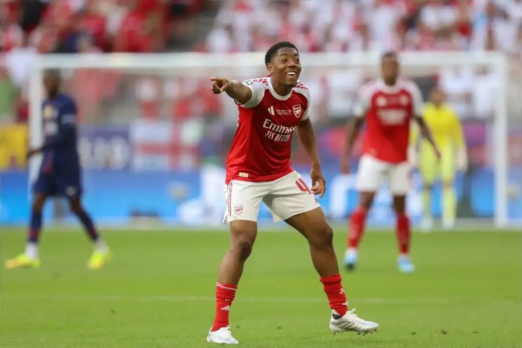 Myles Lewis-Skelly in red Arsenal kit and red socks, smiling and pointing during a match, with blurred players and stadium behind him