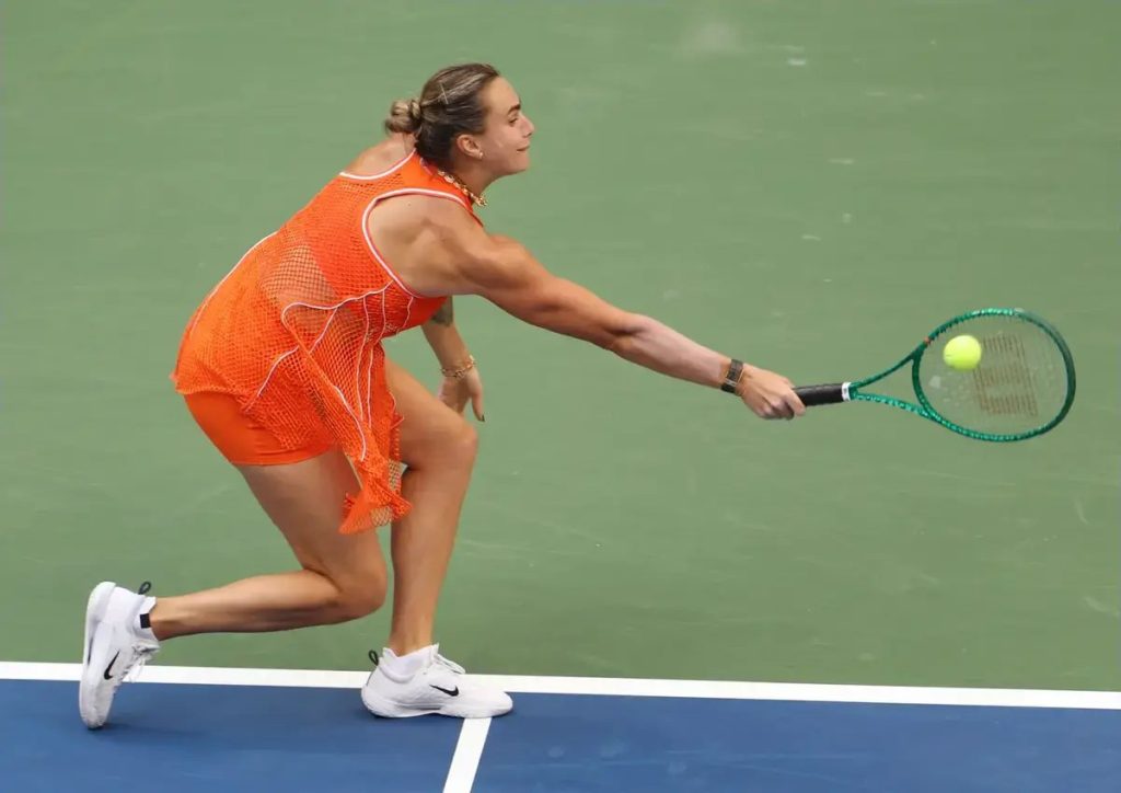 Tennis player in orange mesh outfit lunging to return a ball with a green racket on a hard court at the US Open