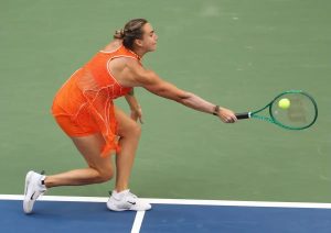 Tennis player in orange mesh outfit lunging to return a ball with a green racket on a hard court at the US Open