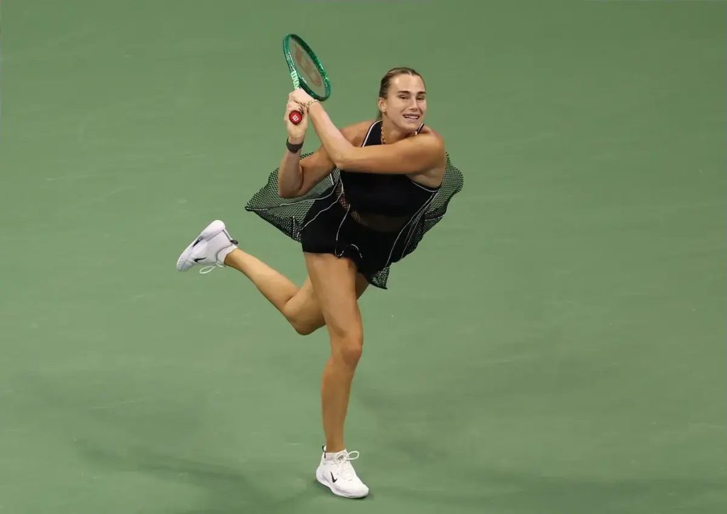 Tennis player in black outfit and green racket mid-stroke on green court, leg raised, at US Open final context