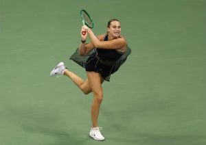 Tennis player in black outfit and green racket mid-stroke on green court, leg raised, at US Open final context