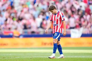 Football player in red-and-white striped Atletico Madrid kit with blue shorts and socks, head down walking on the pitch in a stadium.