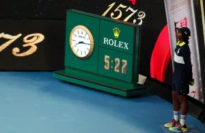 A person in a navy and white Rolex-sponsored outfit stands beside the green Rolex scoreboard showing 5:27 at a tennis court, with a "1573" backdrop.