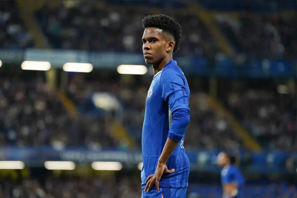 Young footballer in blue Chelsea kit with curly hair, looking over his shoulder in a stadium with blurred crowd behind him