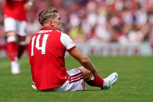 Footballer in red and white Arsenal-style kit, number 14 with name on back, sitting on the pitch holding his leg after a challenge