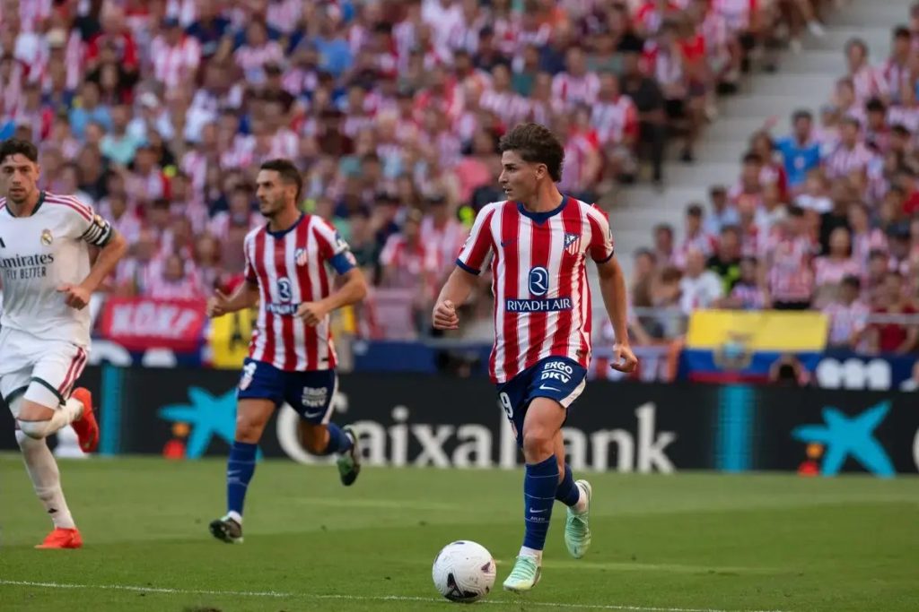 Football player in red-and-white striped Atlético Madrid kit dribbling the ball on the pitch during a match, with a crowd behind him